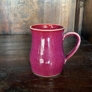 May include: A glossy, deep magenta ceramic mug with a curved, bulbous body and a single side handle. The surface features a speckled, textured finish with a smooth, rounded rim and a consistent, vibrant dark pink color throughout the piece.