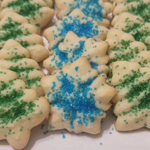 May include: Close-up of Christmas tree-shaped sugar cookies. The cookies are decorated with green and blue sprinkles. The cookies are a light beige color and are arranged closely together.