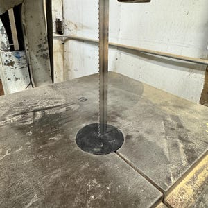 May include: A metal band saw with a blade, set on a dusty, gray work surface. The saw's frame is a mix of gray and off-white, with visible wear. The blade is positioned above a black circular insert.