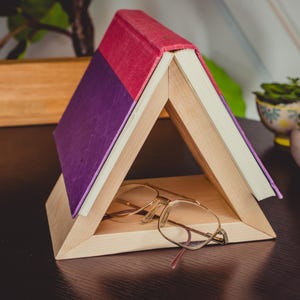 May include: A unique wooden book stand in a triangular shape, holding a book with a purple and red cover. Gold-rimmed eyeglasses rest inside the stand. The stand is on a dark surface, with small potted plants in the background.