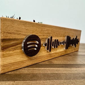 May include: A wooden block with a Spotify logo and a soundwave design. The Spotify logo is a dark brown circle with three curved lines. The soundwave is made of clear acrylic and dark brown accents. The block is made of light brown wood.