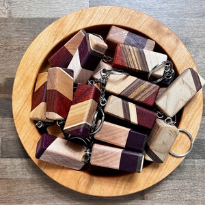 May include: A wooden bowl filled with a variety of colorful wooden keychains. The keychains are made from different types of wood, including maple, walnut, and cherry. Each keychain has a unique grain pattern and color.