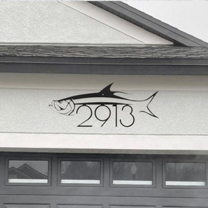 May include: A black silhouette of a fish with the house number 2913 incorporated into the design. The fish is mounted on a white stucco wall.