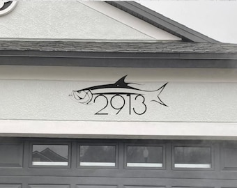 Puede incluir: Una silueta negra de un pez con el número de casa 2913 incorporado en el diseño. El pez está montado en una pared de estuco blanco.