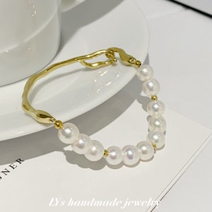 Peut inclure: Un bracelet doré orné de perles blanches. Le bracelet présente un design unique et organique avec une bande texturée et incurvée et un fermoir. Le texte "LY's handmade jewelry" est visible.