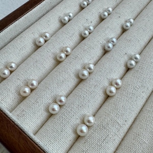 Peut inclure: Une collection de boucles d'oreilles en perles présentées dans un présentoir à bijoux en bois avec une doublure en tissu crème. Les boucles d'oreilles sont disposées par paires, mettant en valeur leur aspect rond et brillant. L'éclairage met en évidence l'iridescence subtile des perles.