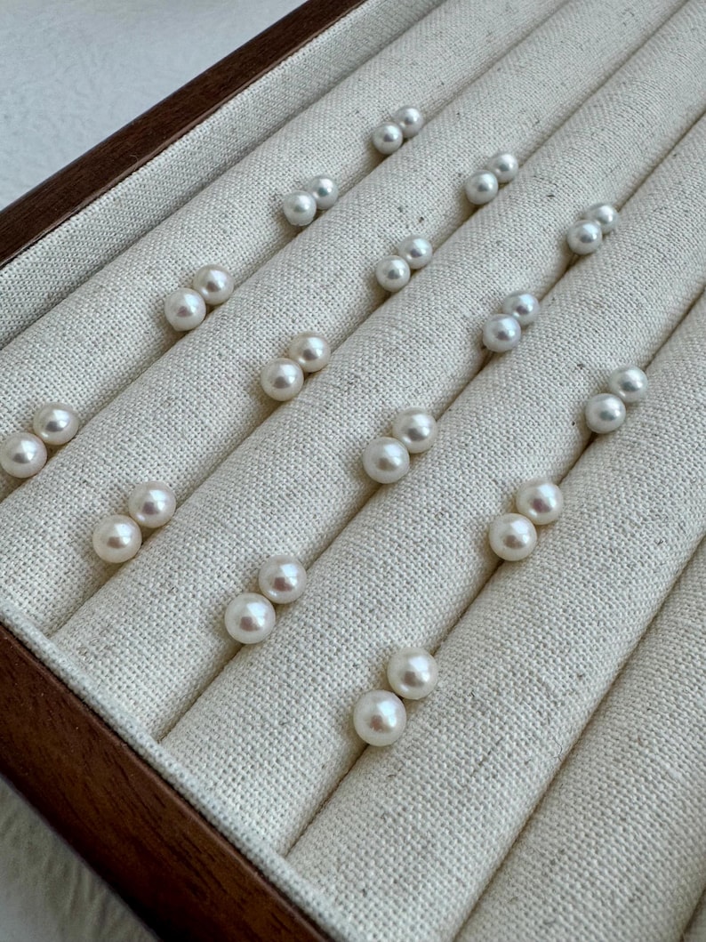 Peut inclure: Une collection de boucles d'oreilles en perles pr&eacute;sent&eacute;es dans un pr&eacute;sentoir &agrave; bijoux en bois avec une doublure en tissu cr&egrave;me. Les boucles d'oreilles sont dispos&eacute;es par paires, mettant en valeur leur aspect rond et brillant. L'&eacute;clairage met en &eacute;vidence l'iridescence subtile des perles.