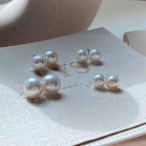 Pode incluir: Tr&ecirc;s pares de brincos de p&eacute;rola branca esf&eacute;rica de tamanhos variados dispostos sobre uma superf&iacute;cie bege clara. Cada brinco apresenta um acabamento liso e lustroso com um pino met&aacute;lico. As p&eacute;rolas exibem reflexos iridescentes subtis.