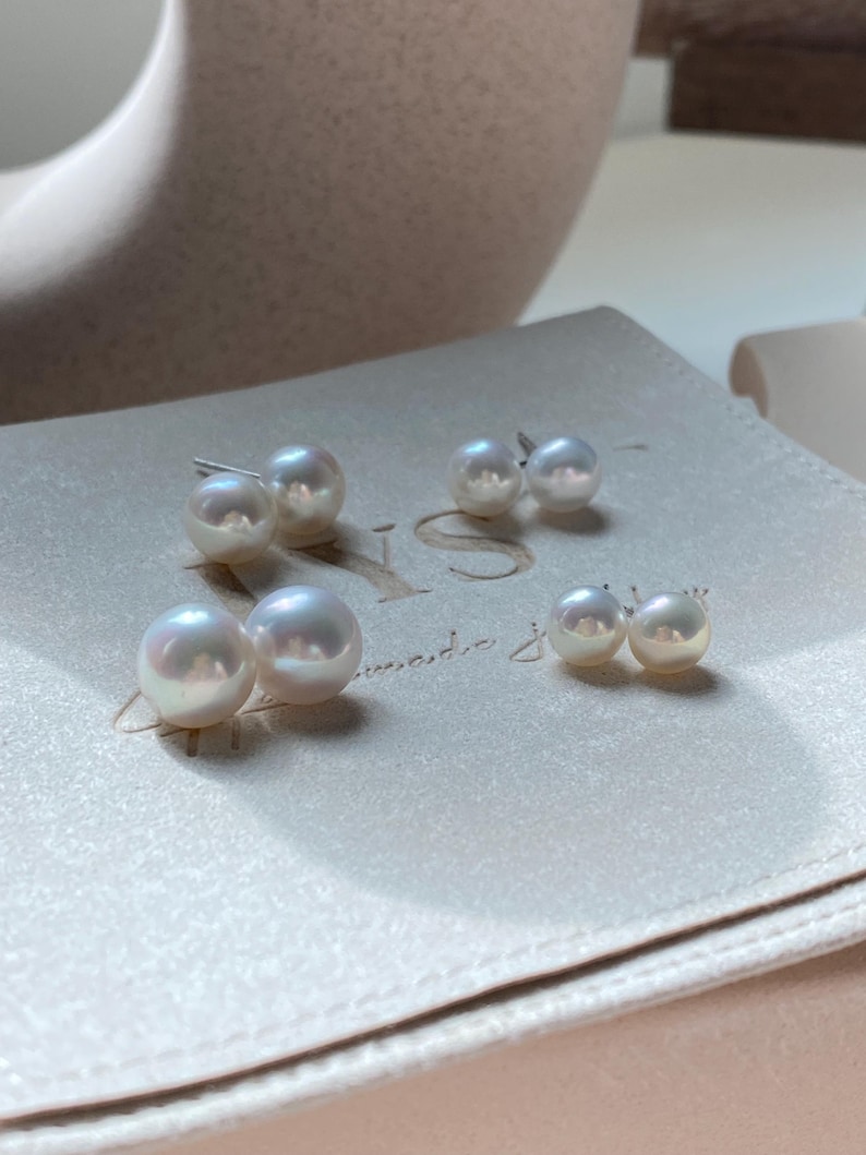 Pode incluir: Tr&ecirc;s pares de brincos de p&eacute;rola branca esf&eacute;rica de tamanhos variados dispostos sobre uma superf&iacute;cie bege clara. Cada brinco apresenta um acabamento liso e lustroso com um pino met&aacute;lico. As p&eacute;rolas exibem reflexos iridescentes subtis.