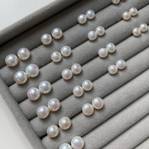 Pode incluir: Uma cole&ccedil;&atilde;o de v&aacute;rios pares de p&eacute;rolas brancas, redondas e brilhantes, dispostas em filas. Cada par consiste em duas p&eacute;rolas esf&eacute;ricas com uma superf&iacute;cie lisa e iridescente, exibidas numa bandeja de joias de veludo cinzento.