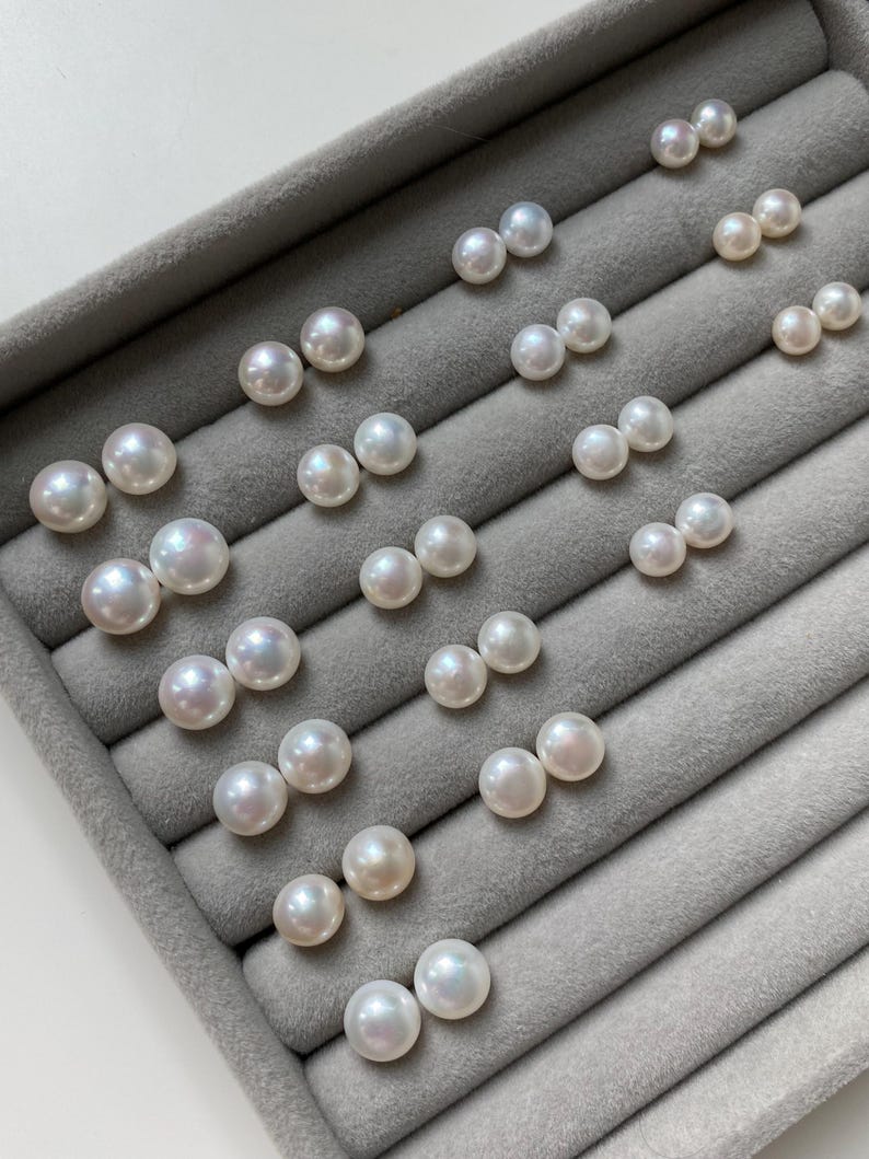 Pode incluir: Uma cole&ccedil;&atilde;o de v&aacute;rios pares de p&eacute;rolas brancas, redondas e brilhantes, dispostas em filas. Cada par consiste em duas p&eacute;rolas esf&eacute;ricas com uma superf&iacute;cie lisa e iridescente, exibidas numa bandeja de joias de veludo cinzento.