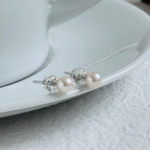 Peut inclure: Deux paires de boucles d'oreilles en perles avec des tiges et des fermoirs en argent sont pr&eacute;sent&eacute;es sur une soucoupe blanche. Les perles sont d'un blanc cass&eacute; cr&eacute;meux, et les boucles d'oreilles sont pos&eacute;es sur un fond blanc.