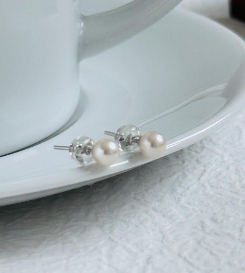 Peut inclure: Deux paires de boucles d'oreilles en perles avec des tiges et des fermoirs en argent sont pr&eacute;sent&eacute;es sur une soucoupe blanche. Les perles sont d'un blanc cass&eacute; cr&eacute;meux, et les boucles d'oreilles sont pos&eacute;es sur un fond blanc.