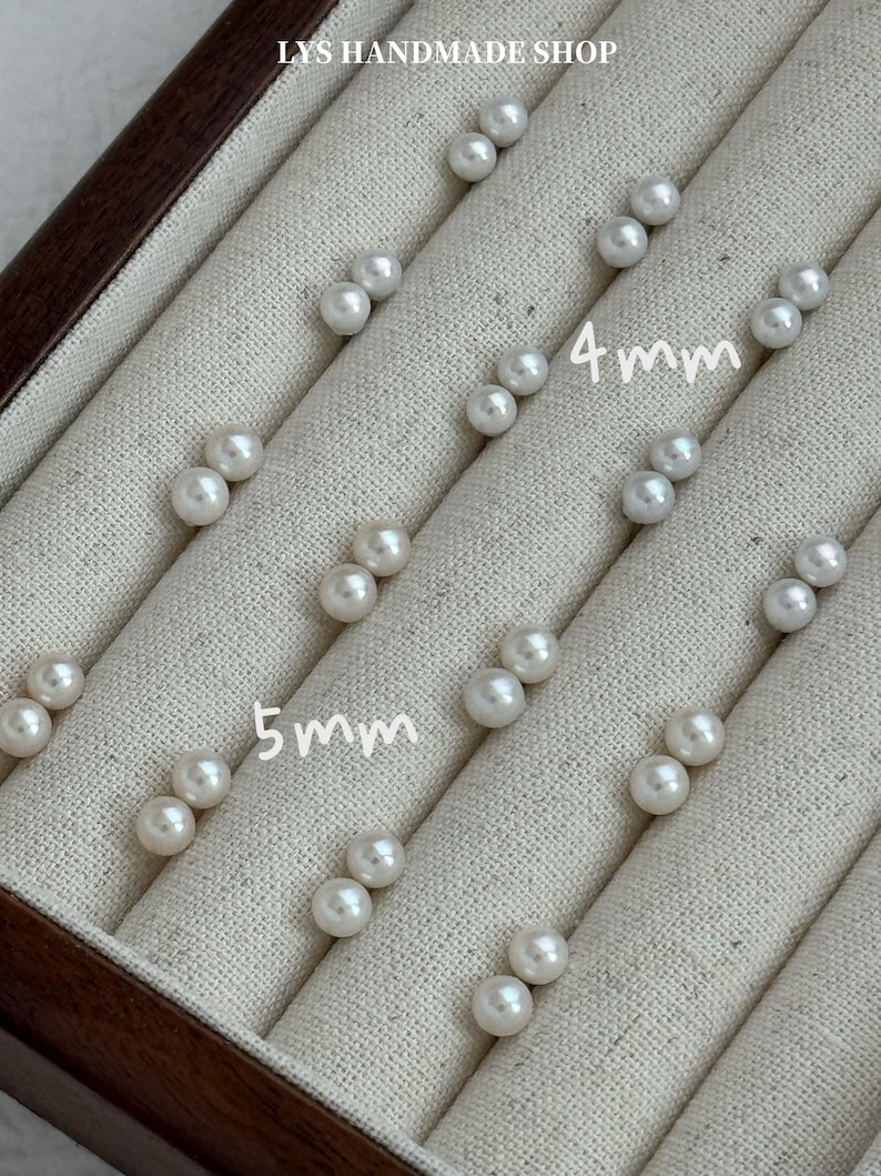Peut inclure: Plusieurs paires de boucles d'oreilles &agrave; clous en perles blanches, certaines de 4 mm et d'autres de 5 mm de diam&egrave;tre, sont pr&eacute;sent&eacute;es dans un &eacute;crin &agrave; bijoux doubl&eacute; de tissu beige. Le texte "LYS HANDMADE SHOP" est visible en haut.
