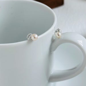 Peut inclure: Une paire de boucles d'oreilles &agrave; clous en perles avec des tiges en argent. Les boucles d'oreilles pr&eacute;sentent de petites perles rondes et claires. Les boucles d'oreilles sont pr&eacute;sent&eacute;es sur le bord d'une tasse en c&eacute;ramique blanche.