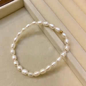 Peut inclure: Un bracelet délicat composé d'une chaîne de perles blanches de forme ovale. Les perles sont enfilées ensemble, avec une petite perle dorée près du fermoir. Le bracelet est présenté sur une surface beige clair.