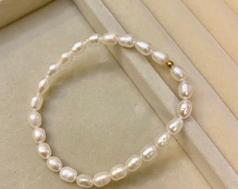 Pode incluir: Uma delicada pulseira com uma fileira de pérolas brancas em forma oval. As pérolas são enfiadas juntas, com uma pequena conta dourada perto do fecho. A pulseira é exibida em uma superfície bege clara.