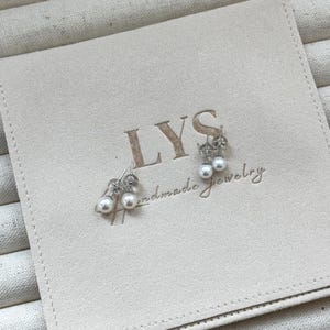 Peut inclure: Deux paires de boucles d'oreilles en perles sur une pochette beige. Les boucles d'oreilles ont de petites perles blanches rondes et des tiges argent&eacute;es. La pochette porte l'inscription "LYS handmade jewelry".