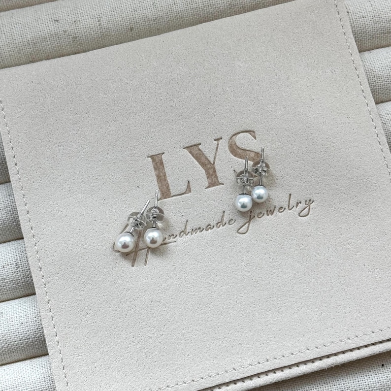 Peut inclure: Deux paires de boucles d'oreilles en perles sur une pochette beige. Les boucles d'oreilles ont de petites perles blanches rondes et des tiges argent&eacute;es. La pochette porte l'inscription "LYS handmade jewelry".