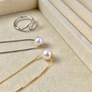 Peut inclure: Une bague en argent au design incurvé unique est posée près de deux boucles d'oreilles en perles avec de fines chaînes. Une boucle d'oreille a une chaîne dorée, l'autre une chaîne argentée. Les perles sont blanches et rondes, sur fond neutre.