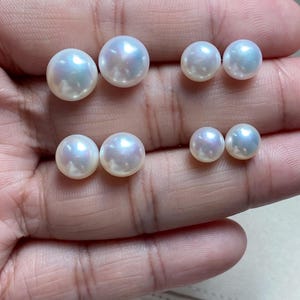 Pode incluir: Quatro pares de p&eacute;rolas redondas, brancas e lustrosas dispostas em duas filas. Cada p&eacute;rola apresenta uma superf&iacute;cie esf&eacute;rica lisa com reflexos iridescentes subtis. As p&eacute;rolas variam em tamanho, com o par maior posicionado no canto superior esquerdo e o par menor no canto inferior direito, exibindo um brilho natural e refletor.