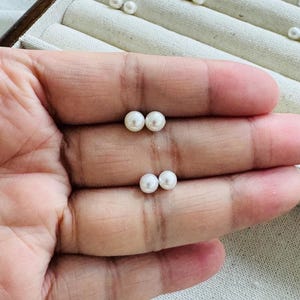 Peut inclure: Deux paires de petites boucles d'oreilles &agrave; clous en perles blanches et rondes sont pr&eacute;sent&eacute;es sur une main. D'autres paires de boucles d'oreilles sont visibles dans une bo&icirc;te &agrave; bijoux en arri&egrave;re-plan. Les perles ont une surface lisse et brillante.