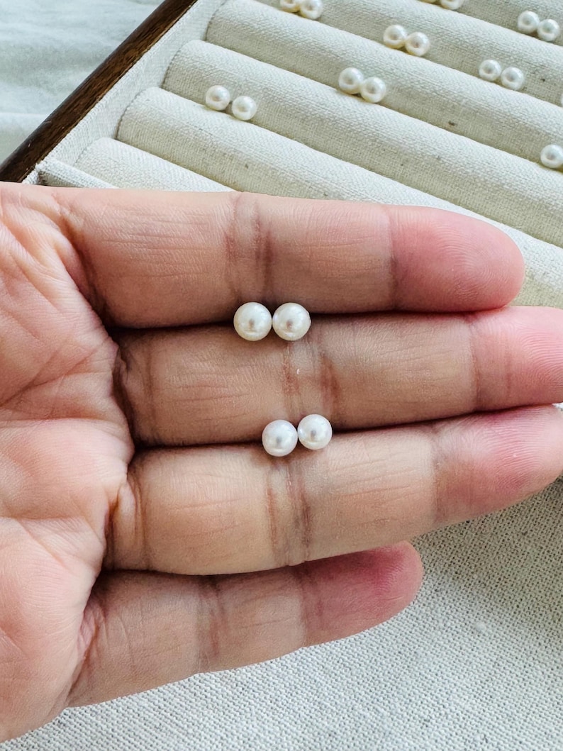 Peut inclure: Deux paires de petites boucles d'oreilles &agrave; clous en perles blanches et rondes sont pr&eacute;sent&eacute;es sur une main. D'autres paires de boucles d'oreilles sont visibles dans une bo&icirc;te &agrave; bijoux en arri&egrave;re-plan. Les perles ont une surface lisse et brillante.