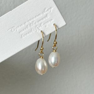 Peut inclure: Boucles d'oreilles dorées avec des perles ovales blanc cassé. Les boucles d'oreilles sont suspendues à des crochets dorés. Une carte blanche avec le texte en relief "Open a warm shop meet the most beautiful you" est en arrière-plan.