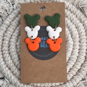 May include: Earrings with a three-tiered design in green, white, and orange, resembling a cartoon character's head. The earrings are attached to a brown card with the text "MAD LOVE DESIGNS". The background is a cream-colored, woven texture.