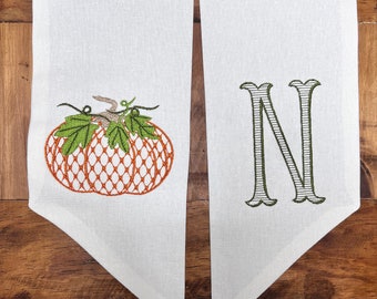 May include: A white fabric table runner with an embroidered pumpkin and the letter "N" in green thread.