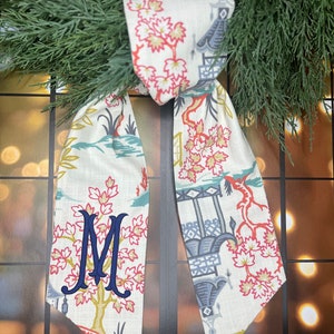 May include: A white fabric bow with a blue and red floral pattern. The bow has a monogram "M" embroidered in navy blue.