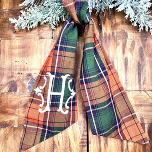 May include: A plaid bow with an embroidered letter "H" in white. The bow is made of orange, green, and brown plaid fabric.