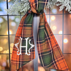 May include: A plaid fabric door swag with a monogrammed "H" in white thread. The swag is in shades of orange, green, and brown.