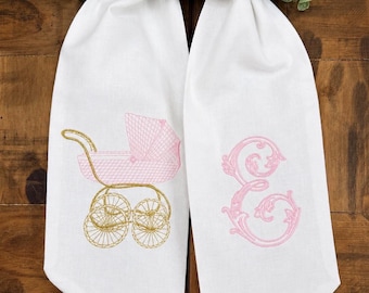May include: A white fabric towel featuring two embroidered designs. On the left, a pink and gold baby carriage with detailed wheels and a handle. On the right, a large, ornate pink letter E in a decorative script font. The fabric is smooth and white.