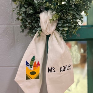 May include: A white fabric bow with a box of crayons and the text "MS. Haile" embroidered on it.
