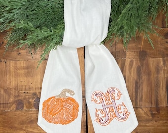 May include: White fabric ribbon with an embroidered pumpkin and the letter "H" in a decorative font. The ribbon is tied around a green wreath.