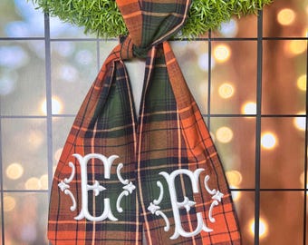 May include: A plaid bow with a white embroidered letter "E" on each side. The bow is tied in a knot and is hanging from a wreath.