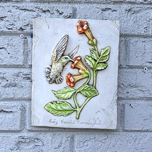May include: A rectangular ceramic wall plaque featuring a raised relief of a ruby-throated hummingbird hovering near orange trumpet-shaped flowers with green leaves. The text "Ruby-throated Hummingbird" is inscribed at the bottom of the piece.
