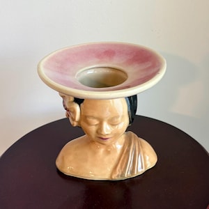 May include: A ceramic vase in the shape of a woman's head and shoulders. The vase has a light skin tone, black hair, and a pink and cream-colored hat-like structure on top. The woman's eyes are closed, and she has a serene expression.