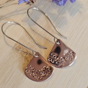 May include: Copper-coloured, fan-shaped earrings with a floral design. The earrings have long, silver-coloured ear wires. They are displayed on a light-coloured wooden surface, with purple flowers in the background.
