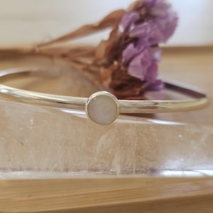 May include: A silver bangle bracelet featuring a central, round, pale stone. The bracelet is presented on a wooden surface, with a dried purple flower in the background. The design is simple and elegant, suitable for everyday wear.
