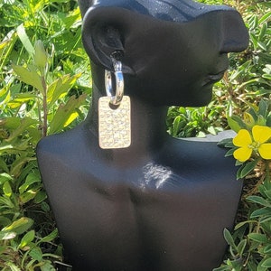 May include: Earrings on a black mannequin head. Each earring has a silver hoop and a gold-coloured rectangular pendant with a textured pattern. The background features green foliage and yellow flowers.