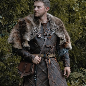 May include: A dark brown leather tunic featuring a diamond-stitched pattern with metal rivets, worn with a thick faux fur shoulder mantle, a chain link chest attachment, and a matching leather satchel bag with a long strap and hanging beads.