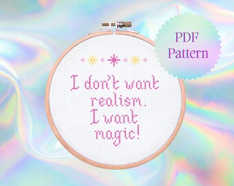 May include: A white embroidery hoop with a pink cross-stitch design that reads "I don't want realism. I want magic!" with yellow stars above the text. The hoop is on a holographic background.