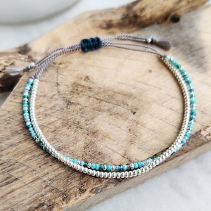 May include: A delicate bracelet with two strands of small, silver-coloured beads and turquoise and teal seed beads. The bracelet is adjustable with a dark teal knot and grey cords. The jewellery rests on a wooden surface.
