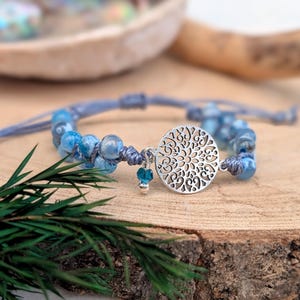 May include: A blue beaded bracelet with a silver charm featuring a detailed openwork design. The bracelet has a sliding knot closure and a small blue bead charm. The bracelet is displayed on a wooden surface.