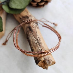 May include: A delicate bracelet made with two strands of small, copper-colored beads. The bracelet is tied with a brown cord and is displayed on a piece of driftwood.
