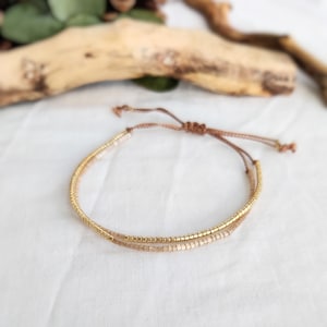 Könnte beinhalten: Ein zartes Armband mit doppelter Schnur aus goldenen und hellbraunen Perlen, gebunden mit einer braunen Schnur.