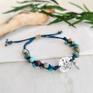 May include: A beaded bracelet featuring turquoise and brown jasper beads, a silver sand dollar charm, and a small shell charm. The bracelet is strung on a dark blue cord with adjustable ties.