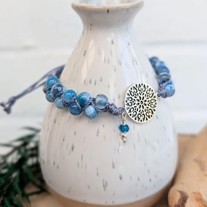 Pode incluir: Uma pulseira de contas azuis com um berloque circular prateado. O berloque apresenta um design vazado detalhado. A pulseira é feita de contas azuis e um cordão azul. Um pequeno berloque azul pende do berloque circular.